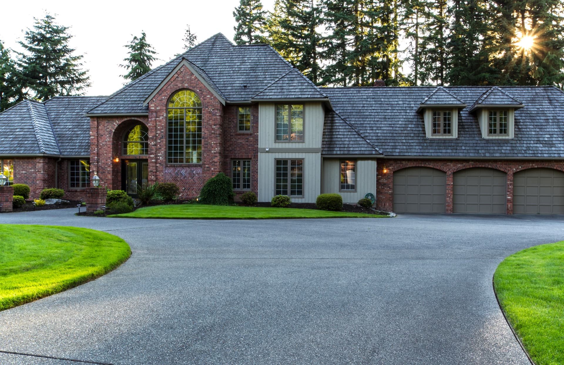 A wide asphalt driveway curving up to the triple garage of a two-storey brick house, with mown lawn either side and tall conifers behind.