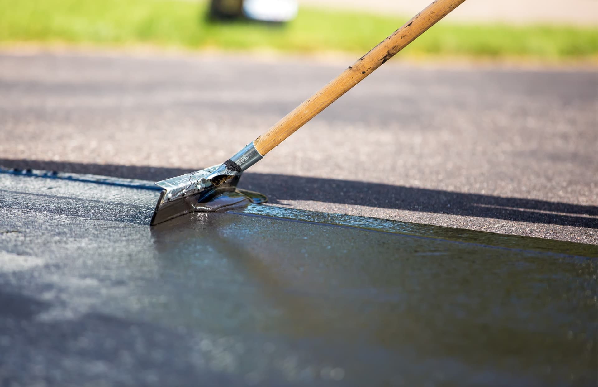 A long-handled squeegee spreading glossy black sealer across grey asphalt in sunlight.